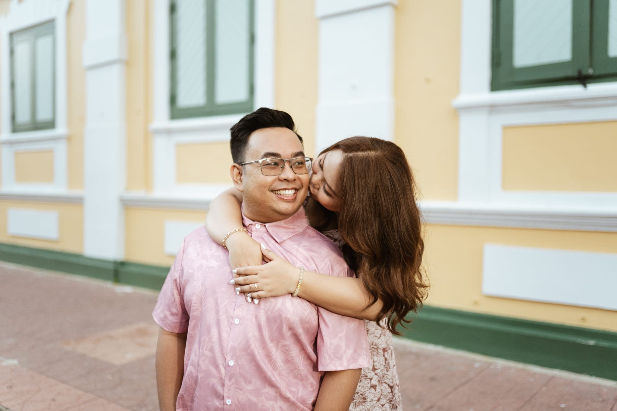 Angela kissing Sam's cheek, her arms around his shoulders, beside a yellow wall with green shutters