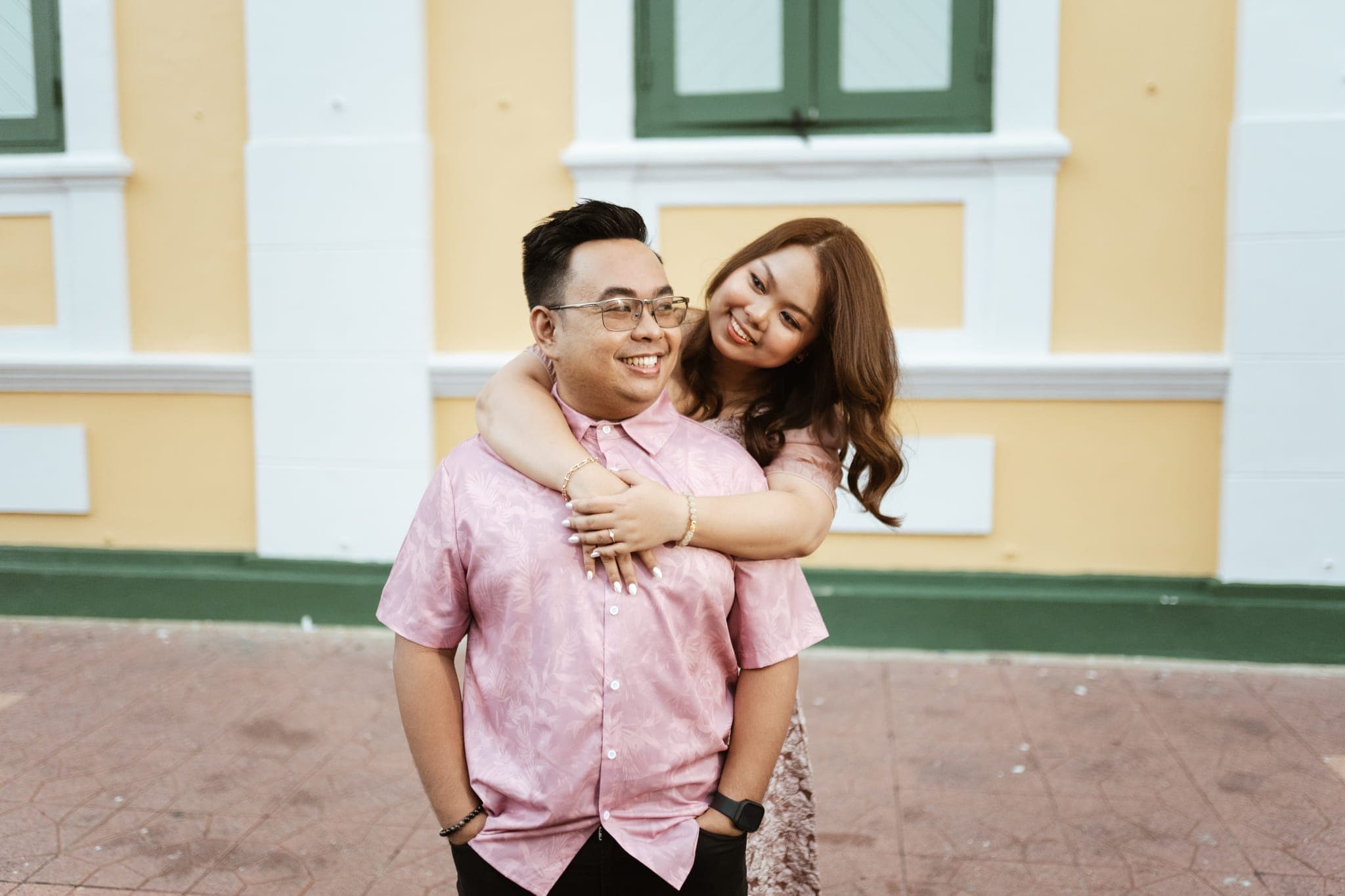 Angela hugging Sam from behind as both smile beside a yellow-and-white wall