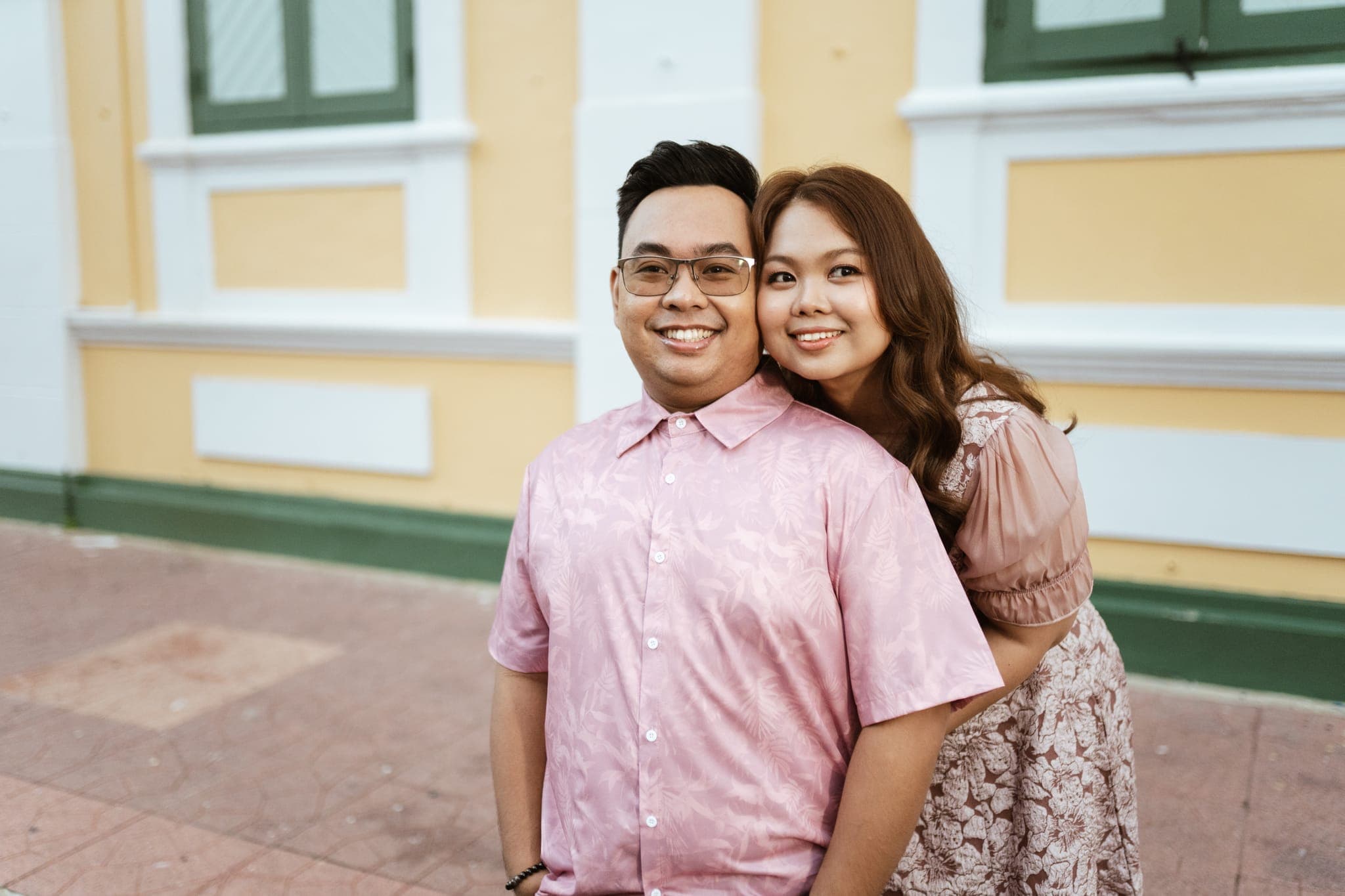 Sam and Angela smiling side by side in front of a yellow-and-white wall