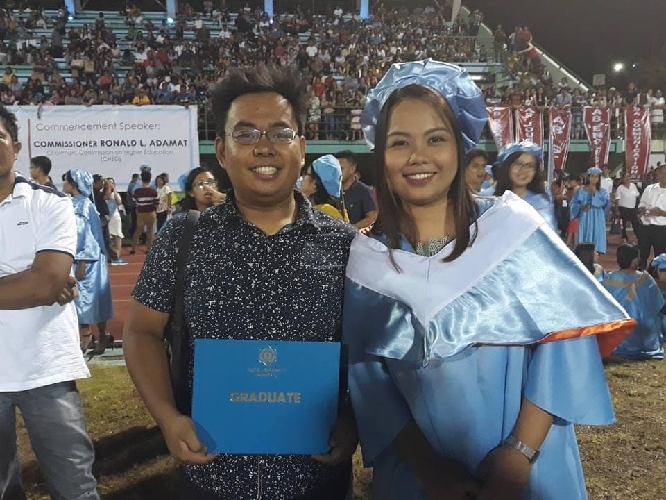 Sam and Angela at a graduation ceremony; Angela wears a pale blue toga and Sam holds a blue diploma folder