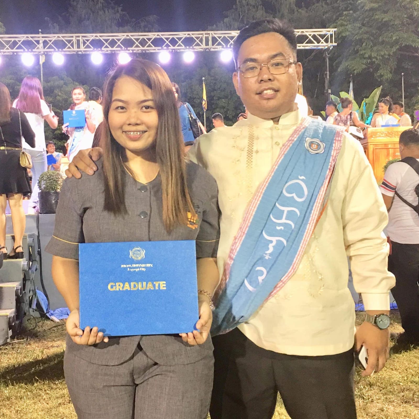 Sam and Angela at an evening graduation; Sam wears a sash over a barong and Angela holds a blue diploma folder