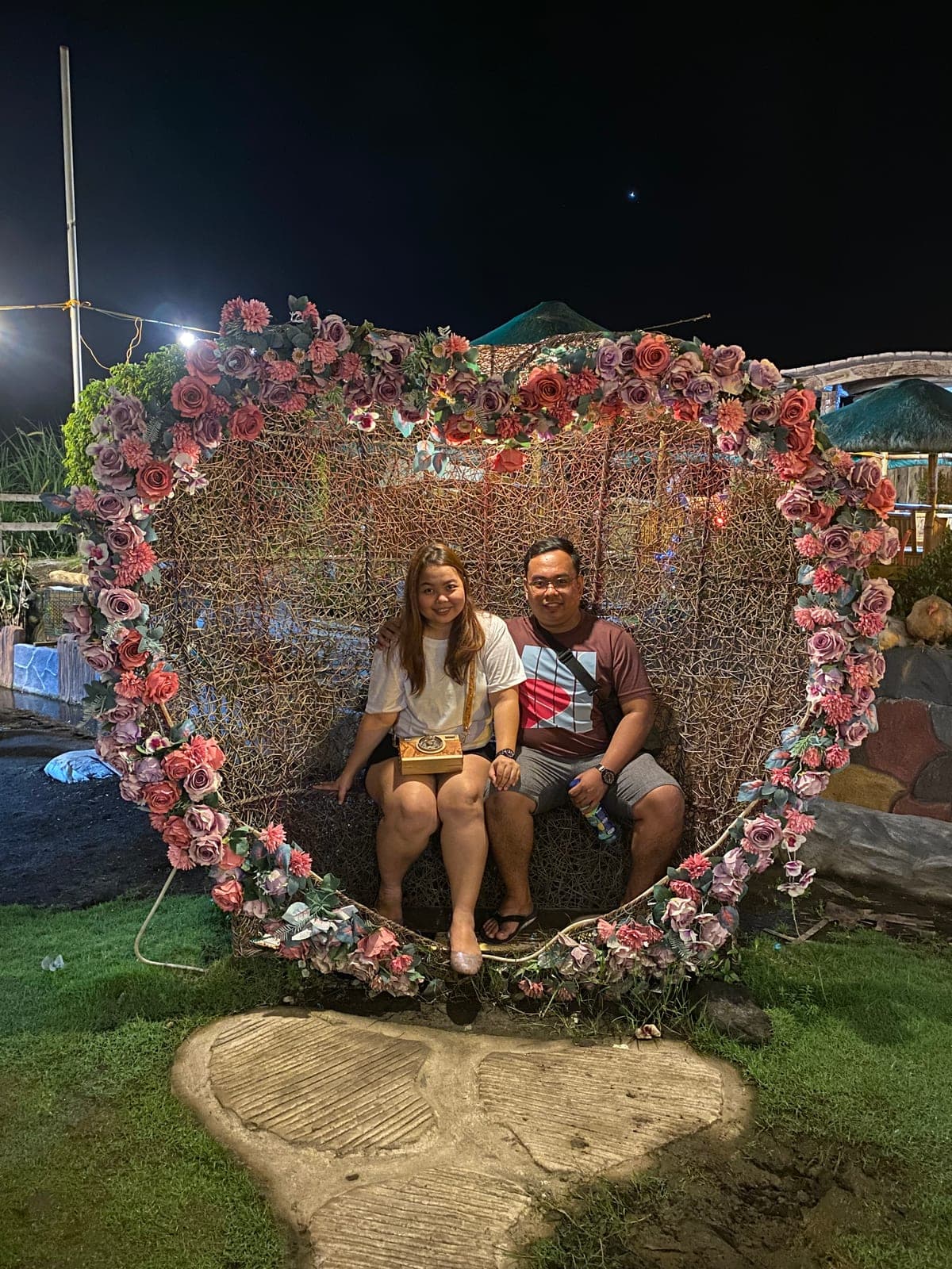 Sam and Angela sitting inside a large heart-shaped flower arch at night
